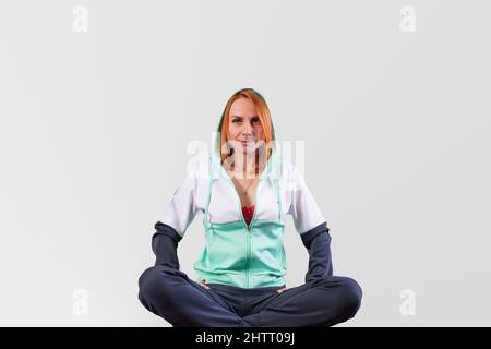 Ein süßes Mädchen, in einem Trainingsanzug, sitzt auf dem Boden, die Beine gekreuzt, auf einem grauen Hintergrund. Hochwertige Fotos Stockfoto