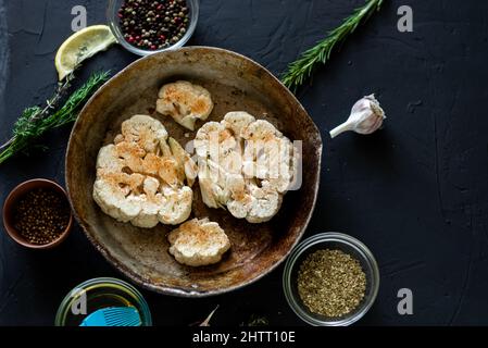 Blumenkohlsteak Kochen. Roher Blumenkohl, mit Gewürzen bestreut, liegt in einer Bratpfanne. Olivenöl, Kräuter, verschiedene Gewürze in der Nähe. Dunkler Hintergrund. Stockfoto