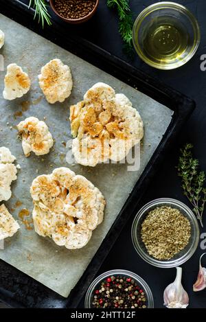 Blumenkohlsteak Kochen. Roher Blumenkohl, mit Gewürzen bestreut, liegt auf einem Backblech. Olivenöl, Kräuter, verschiedene Gewürze in der Nähe. Dunkler Hintergrund. Stockfoto