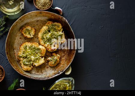 Blumenkohlesteak mit Gewürzen liegt in einer Pfanne. Olivenöl, Chimichurri-Sauce, Kräuter, verschiedene Gewürze nebeneinander. Dunkler Hintergrund. Für Text platzieren. Stockfoto