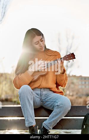 Junge Latina spielt Ukulele in einem Stadtpark. Frau sitzt auf einer Bank und übt mit ihrem Musikinstrument. Stockfoto