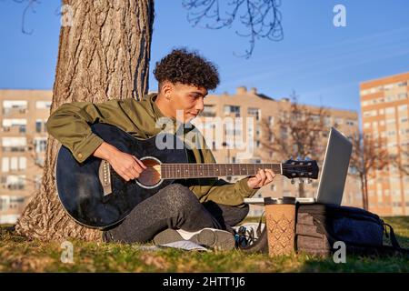 Autodidaktischer junger Mann, der mit dem Laptop Gitarre spielt und lernt. Teenager, der ein Lied komponiert. Stockfoto