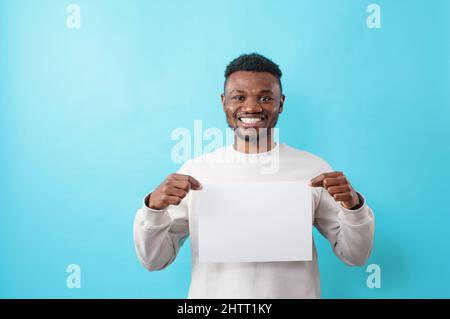 Ein junger, lächelnder Afroamerikaner hält ein weißes Blatt in den Händen, auf einem blau isolierten Hintergrund Stockfoto