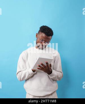 Ein junger afroamerikanischer Mann mit Brille schreibt Informationen auf blauem Hintergrund in ein Notizbuch Stockfoto