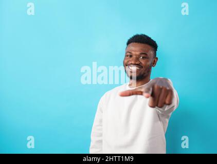 Ein junger afroamerikanischer Mann, der in legerer Kleidung gekleidet ist, lächelt fröhlich und zeigt seinen Finger auf blauem Hintergrund positiv nach vorne. Stockfoto