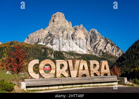 Mountain Mt. Sassongher, Sass Songher, ragen über der Stadt und bunten Bäumen im Herbst, der Name Corvara in Holz geschnitten. Stockfoto