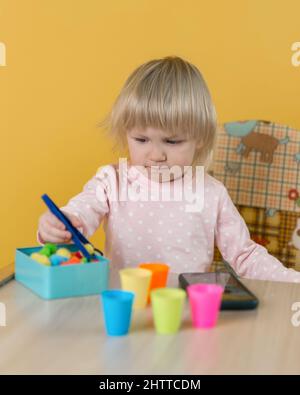 Ein zweijähriges Mädchen sitzt an einem Tisch gegen eine gelbe Wand und legt Kugeln in bunten Cups nach Farben aus Stockfoto