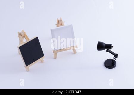 Zwei hölzerne Easels Miniatur mit blanker schwarzer und weißer quadratischer Leinwand für Künstler und Maler - Mockup. Mini-Holzständer mit sauberer Tafel und kleiner schwarzer Tischleuchte auf weißem Hintergrund, Kopierraum Stockfoto