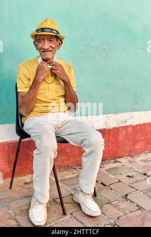 Älterer Einheimischer mit gelbem Hut sitzt auf einem Stuhl in der Nähe einer aquamarinfarbenen Wand in Trinidad, Kuba. Stockfoto