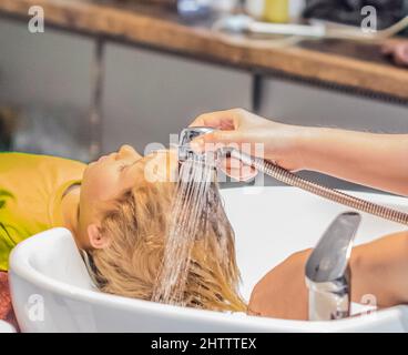 Wet Head Blonde Little Boy liegt auf dem Waschbecken. Weibliche Hand. Haarwaschmassage bekommen Shampoo Schaum, Dusche Wasserstrahl. Schönheitssalon Stockfoto