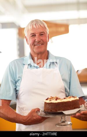 Etwas, um den süßen Zahn zu befriedigen. Porträt eines glücklichen älteren Mannes, der einen frisch gebackenen Kuchen in seiner Bäckerei hält. Stockfoto