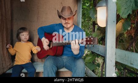 Papa Cowboy spielt für sein kleines Kind eine akustische Gitarre. Altes Retro-Dekor Stockfoto