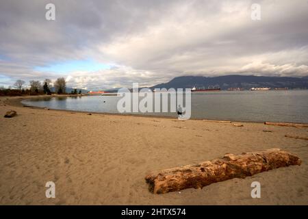 Frau, die im Winter mit der English Bay mit Frachtern im Hintergrund am Strand von New York entlang läuft, Vancouver, British Columbia, Kanada