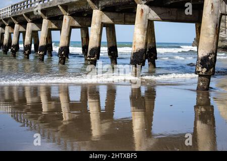 Lange, sich wiederholende Strukturmuster, Reflexion im nassen Sand und abnehmende Perspektive und Schatten der Struktur unter Tolaga Bay Wharf, Ostküste N Stockfoto