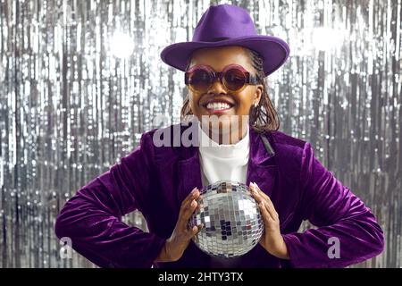 Positive junge dunkelhäutige Frau im Samtanzug, die mit beiden Händen den Diskoball hält. Stockfoto