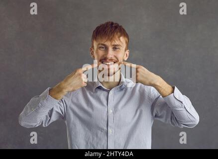 Junger Mann in einem legeren Hemd mit weißen Zähnen, die auf grauem Hintergrund stehen Stockfoto