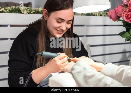 Master Professional in Schönheitssalon in schwarzer Uniform mit elektrischen Nagelfeile Bohrer macht Maniküre zu einer Frau Client. Stockfoto