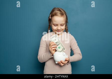 Hübsches Mädchen, das eine Dollar-Banknote auf blauem Hintergrund in ein Sparschwein legt Stockfoto