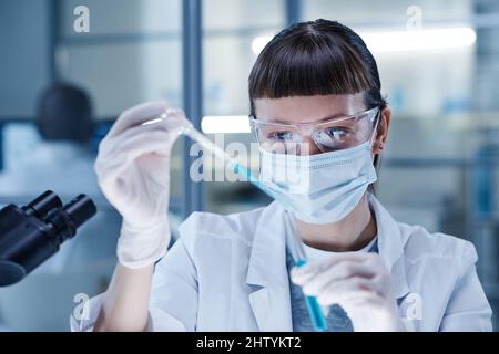 Junge Frau in Maske goss die blaue Flüssigkeit während der medizinischen Forschung im Labor in das Reagenzglas Stockfoto