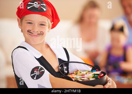 Ein Korb voller Leckereien. Ein kleiner Junge, der in einem Piratenkostüm gekleidet ist und einen Korb mit Süßigkeiten in der Hand hält, während die Familie im Hintergrund sitzt. Stockfoto