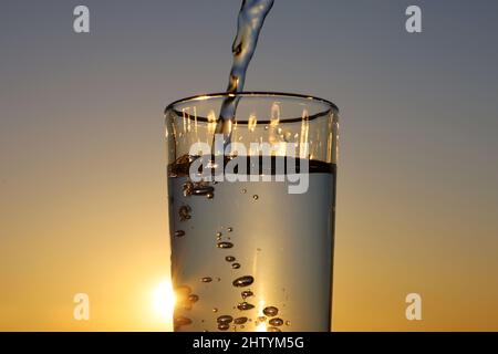 Sauberes Wasser, das auf dem Hintergrund des Sonnenuntergangs in das Trinkglas fließt. Konzept von Gesundheit und Frische, Durst, Wasserreinigung Stockfoto