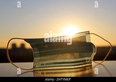 Medizinische Maske auf dem Hintergrund von strahlender Sonne und Stadtgebäuden bei Sonnenuntergang. Konzept der Sicherheitsmaßnahmen während der Coronavirus-Pandemie Covid-19 Stockfoto