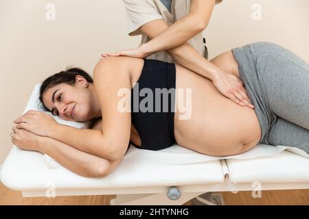 Physiotherapeutin, die eine auf der Seite einer Schwangeren auf einem Massageliegen in einem Gesundheitszentrum liegende Frau massiert Stockfoto