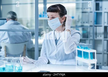 Labortechniker in Maske sitzt im Labor am Tisch und arbeitet mit Proben in Reagenzgläsern Stockfoto