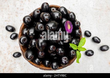 Schwarze reife Syzygium cumini Früchte. Dunkelschwarze java-Pflaume in einer Holzschale auf isoliertem weißem Hintergrund. Grünes Minzblatt auf einigen großen java-Pflaumen. Stockfoto