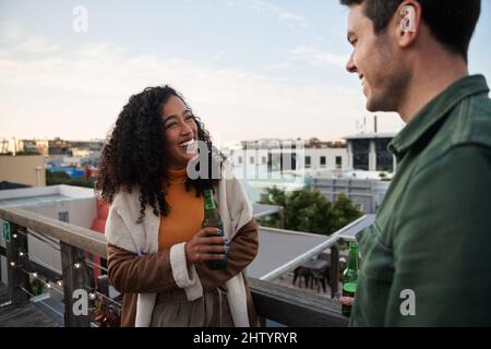 Biracial junge Erwachsene Frau lachen mit einem Freund auf dem Balkon einer Dachterrasse in der Stadt. Trinken Sie in der Hand, Blick auf die Stadt Stockfoto