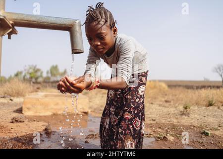 Hübsches kleines schwarzes Mädchen, das von einer westafrikanischen Dorfpumpe überkrümmt wird und glücklich beide Hände hält, um das aus dem Rohr fliessende frische Wasser zu fangen; wat Stockfoto