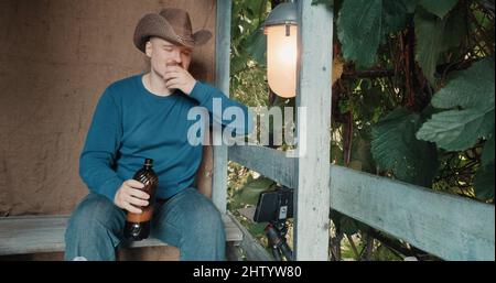 Cowboy-Mann auf der Veranda trinkt Bier aus einer Flasche und kommuniziert online per Telefon Stockfoto