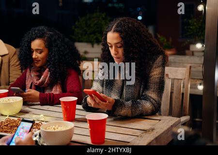 Zwei Frauen mit birazialem jungen Erwachsenen, die auf einer Party sitzen und eine SMS an ihr Telefon senden. Antisozial, mit Technologie auf der Dachparty Stockfoto