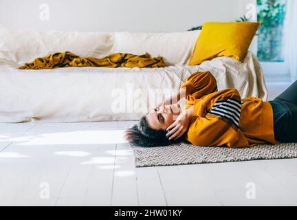 Frau in legerer Kleidung mit Kopfhörern, die in ihrem Wohnzimmer auf dem Boden liegt und gerne Musik hört. Freizeit der alleinstehenden Frau entspannend Stockfoto