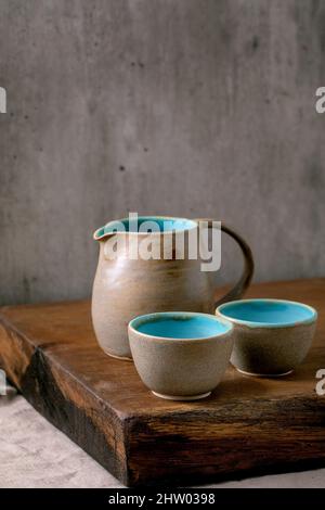 Set aus leerem türkis-grauen strukturierten Keramikkrug und Tassen, die auf einem Holztisch mit grauer Wand im Hintergrund stehen Stockfoto