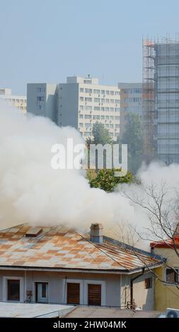 Ansicht der Feuerwehrleute, die das Feuer aus dem brennenden Haus mit Wasser löschen. Rauch und Flammen, die durch ein Feuer vom Dach des Gebäudes entstehen und gefährlichen Smog und Rauch in der Nachbarschaft der Stadt ausbreiten. Stockfoto