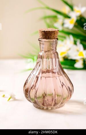 Schöne rosa transparente Flasche Parfüm mit Kork, auf weißem Marmortisch stehend. Kleine weiße Blüten, Frühlings-Narzissen im Hintergrund. Parfüm m Stockfoto