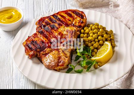 Saftige gegrillte Schweinefleisch-T-Bone-Steaks mit gekochten grünen Erbsen und Zitrone auf einem weißen Teller auf einem Holztisch, horizontale Ansicht von oben Stockfoto