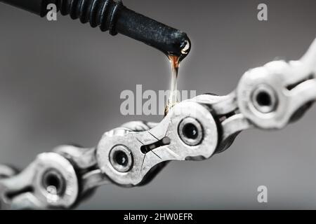 Schmieren einer Fahrradkette mit einem Tropfen goldenem Öl aus nächster Nähe auf grauem Hintergrund. Pflege des Fahrradantriebs. Stockfoto