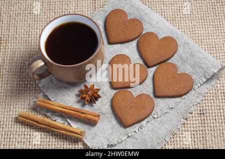 Herzförmige Ingwerkekse, Tasse Kaffee und Gewürz auf rauem Stoffhintergrund Stockfoto