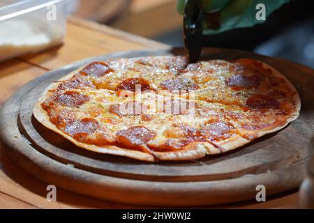 Eine Person mit grünen Handschuhen schneidet dünnkruste, kleine Pizzen mit einem Pizzabschneider auf einem Holztablett. Stockfoto