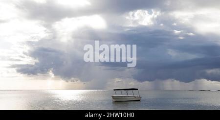 Ein Boot schwimmt vor der Südwestküste von Mauritius. Sonnenlicht durchbricht die launischen Wolken über dem Indischen Ozean. Stockfoto