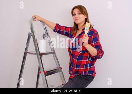 Eine Malerin im karierten Hemd steht auf einer Leiter mit einem Pinsel in der Hand Stockfoto