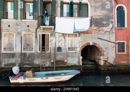 Einheimische Frau auf Balkon über dem Wasserkanal in Venedig, mit einem Boot vor Anker, parkt am Eingang zum Haus, Wäschetrocknung auf dem Balkon, Italien Stockfoto