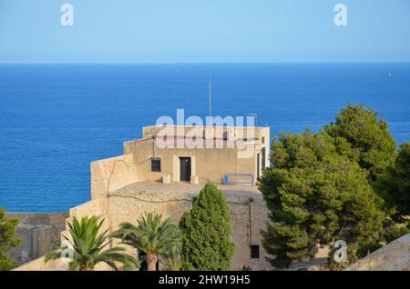 Alicante, Spanien - Juli 17 2019: Einige Burggebäude hinter grünen Palmen aus dem Castillo de Santa Bárbara in Alicante unter blauem, sonnigem Himmel. Im Stockfoto