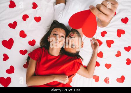 Draufsicht auf ein wunderschönes junges verliebtes Paar, das auf einem weißen Bett mit roten Herzen liegt. Liebe und Beziehungen Stockfoto