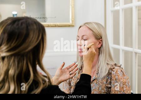 Kosmetikerin, die Foundation Make-up auf eine blonde Frau in einem Schminkstuhl anwendet Stockfoto