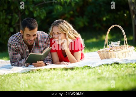 Paare, die zusammen lachen Aufnahme eines glücklichen jungen Paares, das ein Buch liest, während er ein Picknick in einem Park macht. Stockfoto