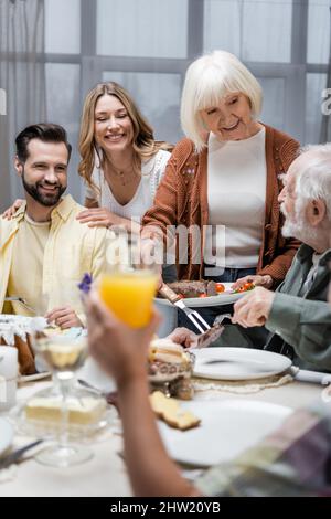 Lächelnde ältere Frau serviert gebratenes Fleisch in der Nähe der Familie und verschwommenes Kind mit einem Glas Orangensaft, Stockbild Stockfoto