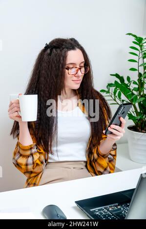 Schöne Frau in Gläsern trinkt Kaffee in einer Pause und mit dem Smartphone am Arbeitsplatz. Frau, die zu Hause arbeitet, eine Schulung absolviert, ein Webinar ansieht, o Stockfoto
